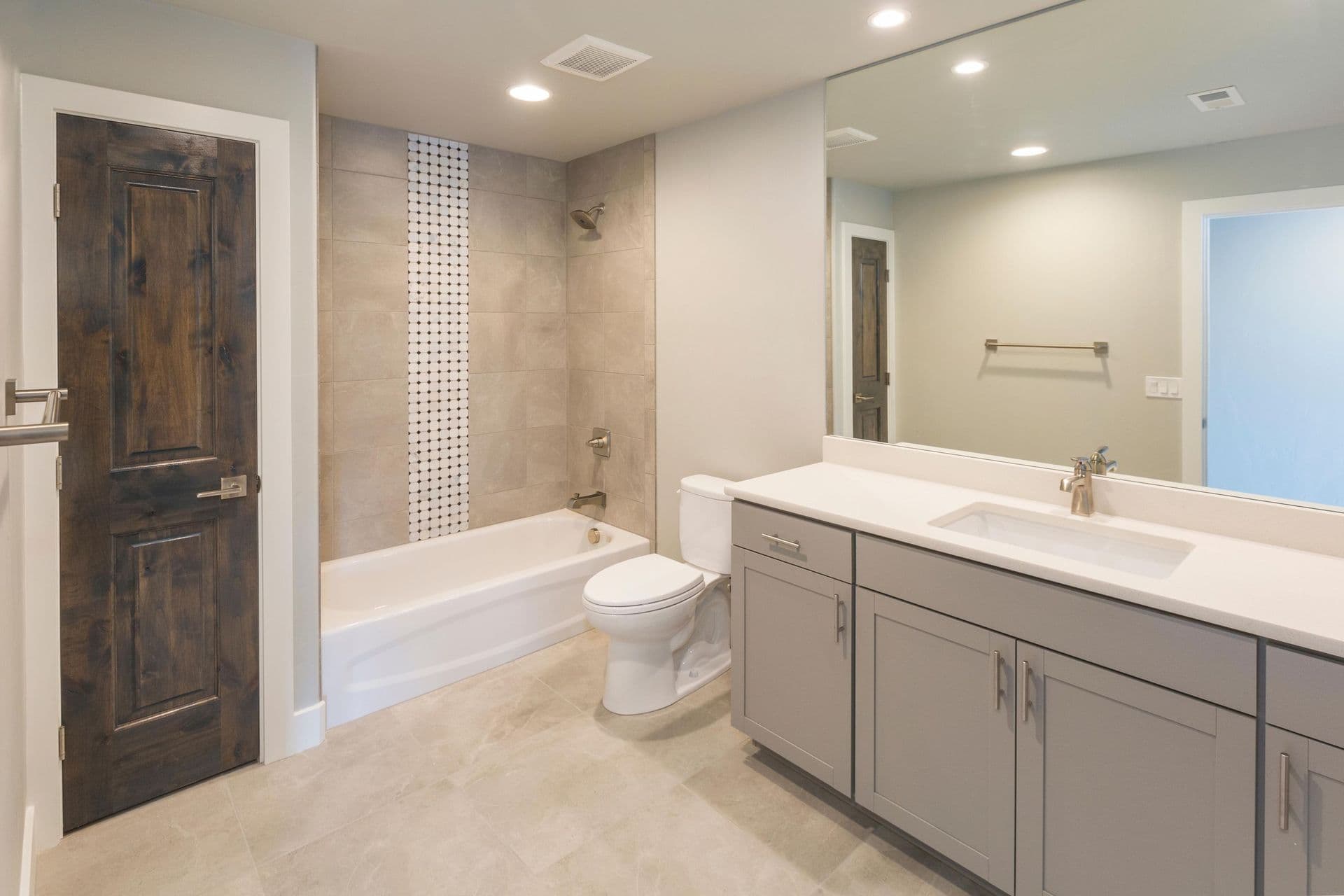 A light gray bathroom with tan marbled tile floor and combination shower bathtub with black and white tile accent feature.