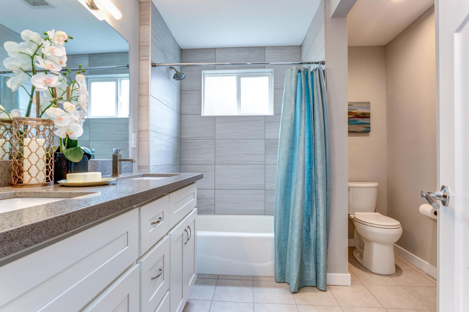 Bright bathroom with modern decor, with a granite countertop with a floral arrangement, white cabinets, a bathtub with blue curtain, and a toilet.
