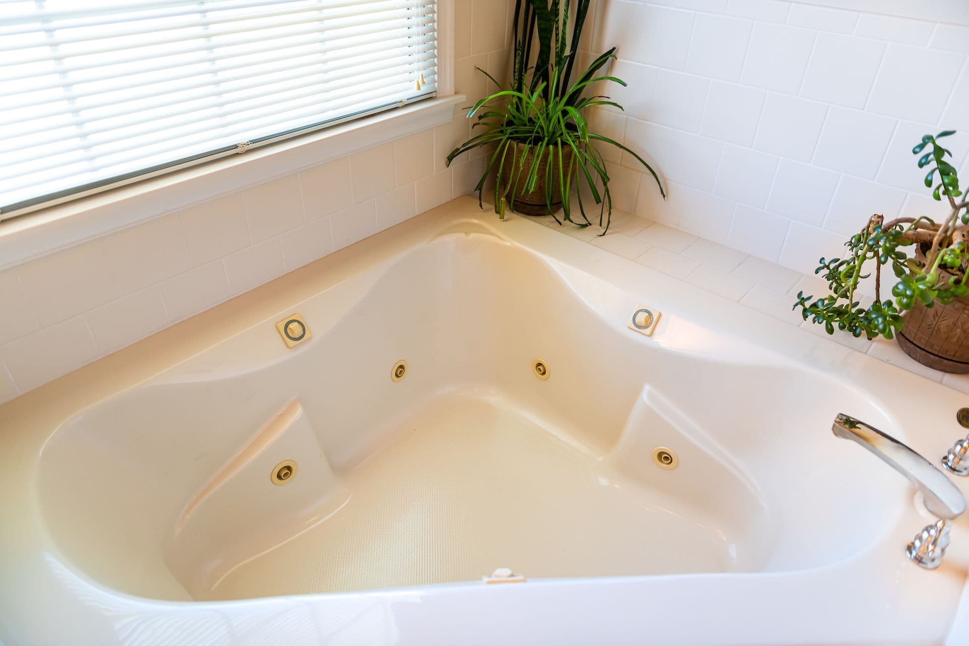 Corner bathtub with jets, surrounded by white tiles, houseplants, and a window with blinds.
