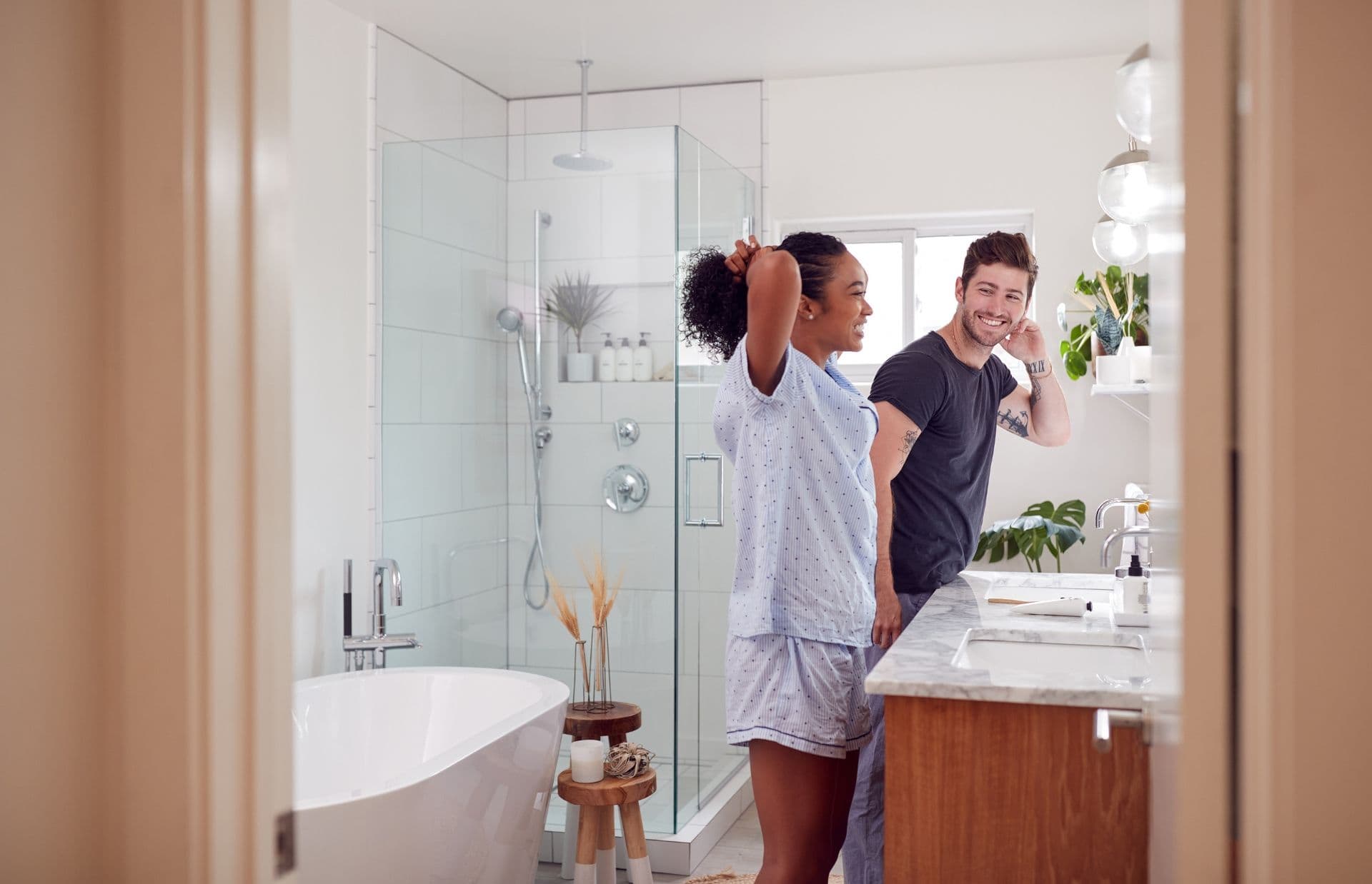 A couple in a modern bathroom, both smiling, one in pajamas, the other in casual clothes. Bright, airy space with plants and glass shower.