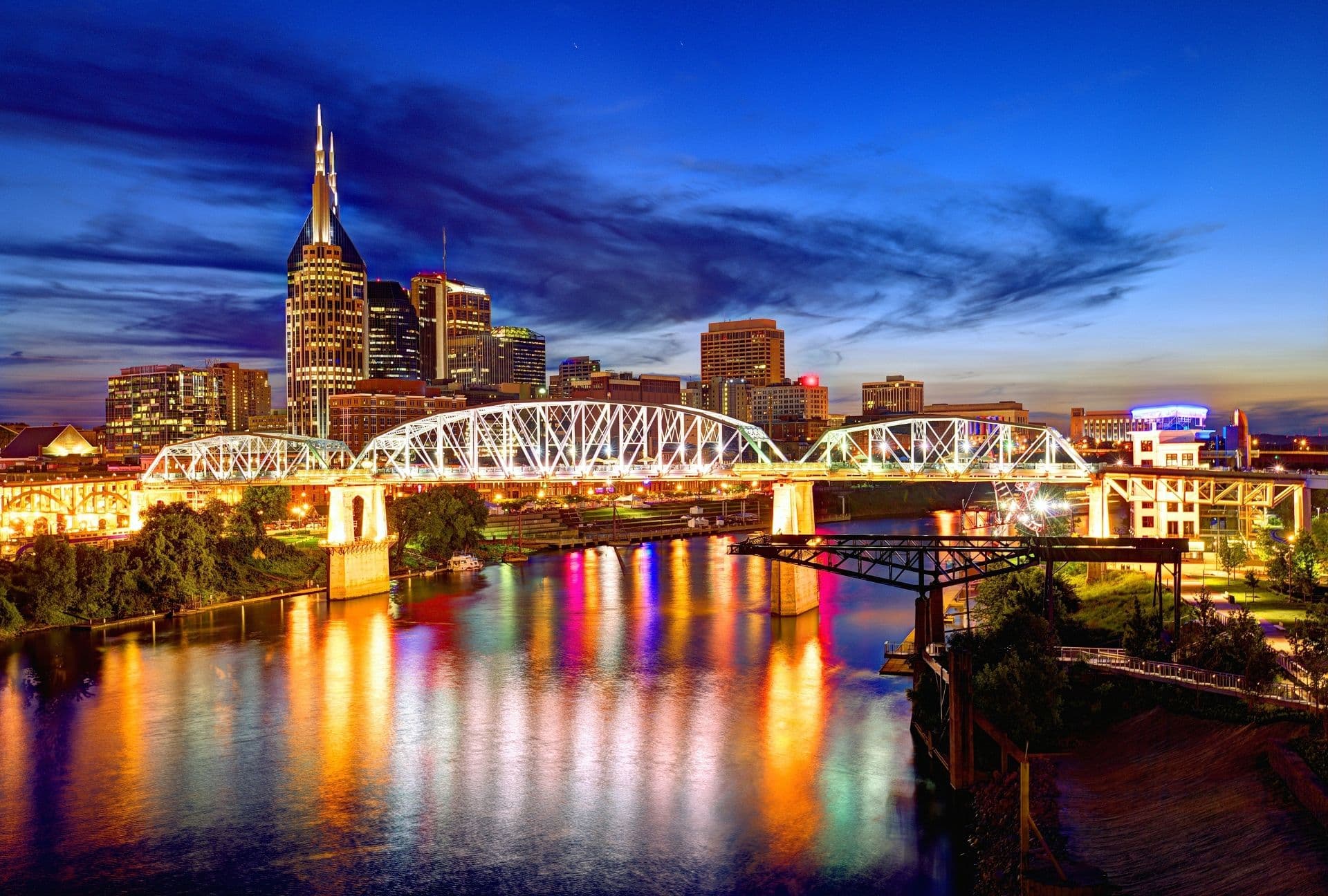 Cityscape of Nashville at twilight with a lit-up bridge spanning a river. The skyline features illuminated skyscrapers, reflecting vibrant colors on the water.