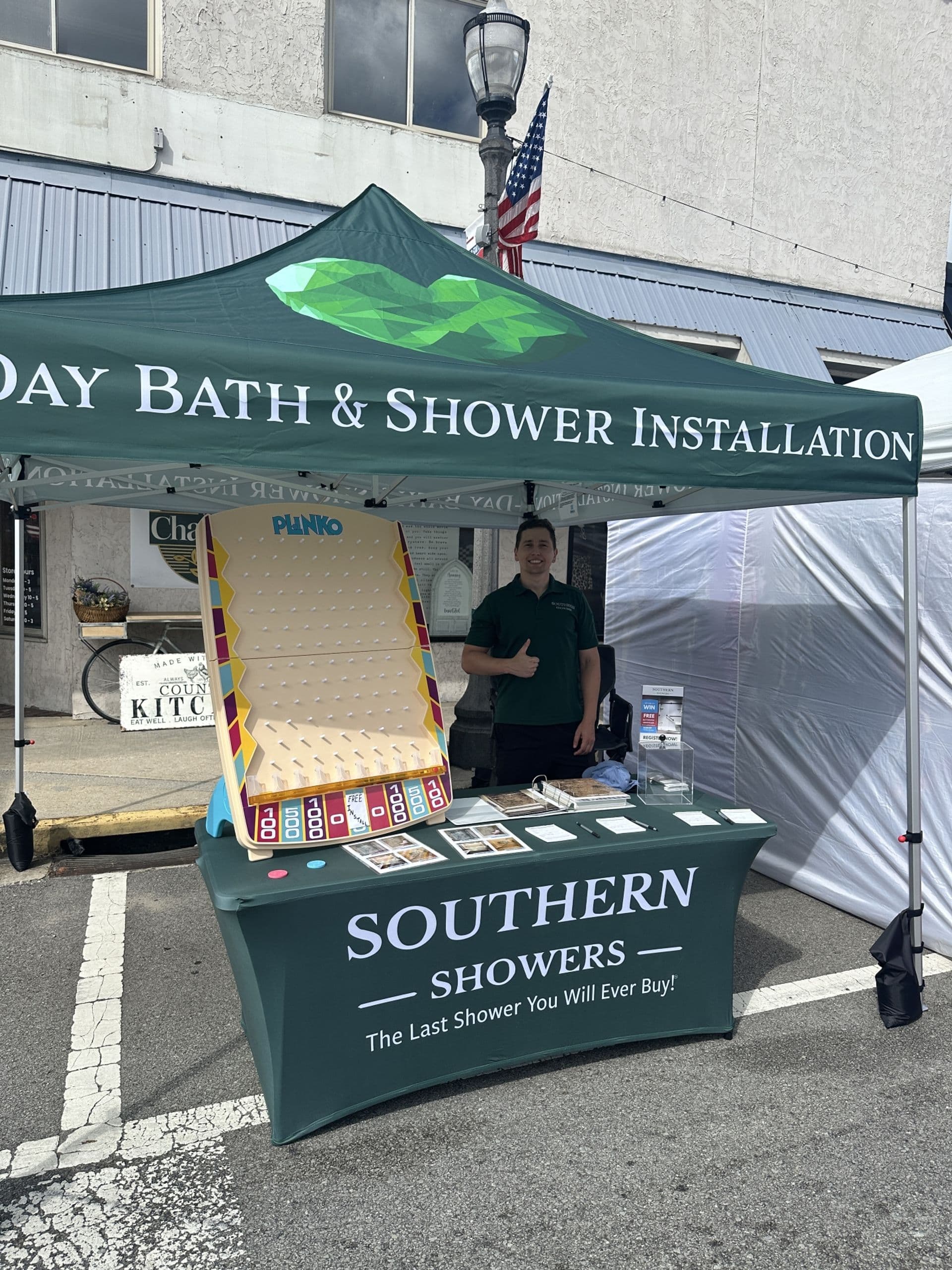 A booth with a green canopy labeled "Bath & Shower Installation." A person stands behind a table that has brochures and a Plinko game.
