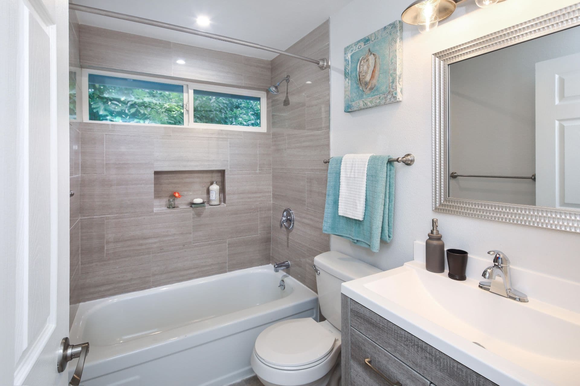 A bright, modern bathroom with beige tiles. Features a bathtub, vanity with a mirror, and turquoise accents.