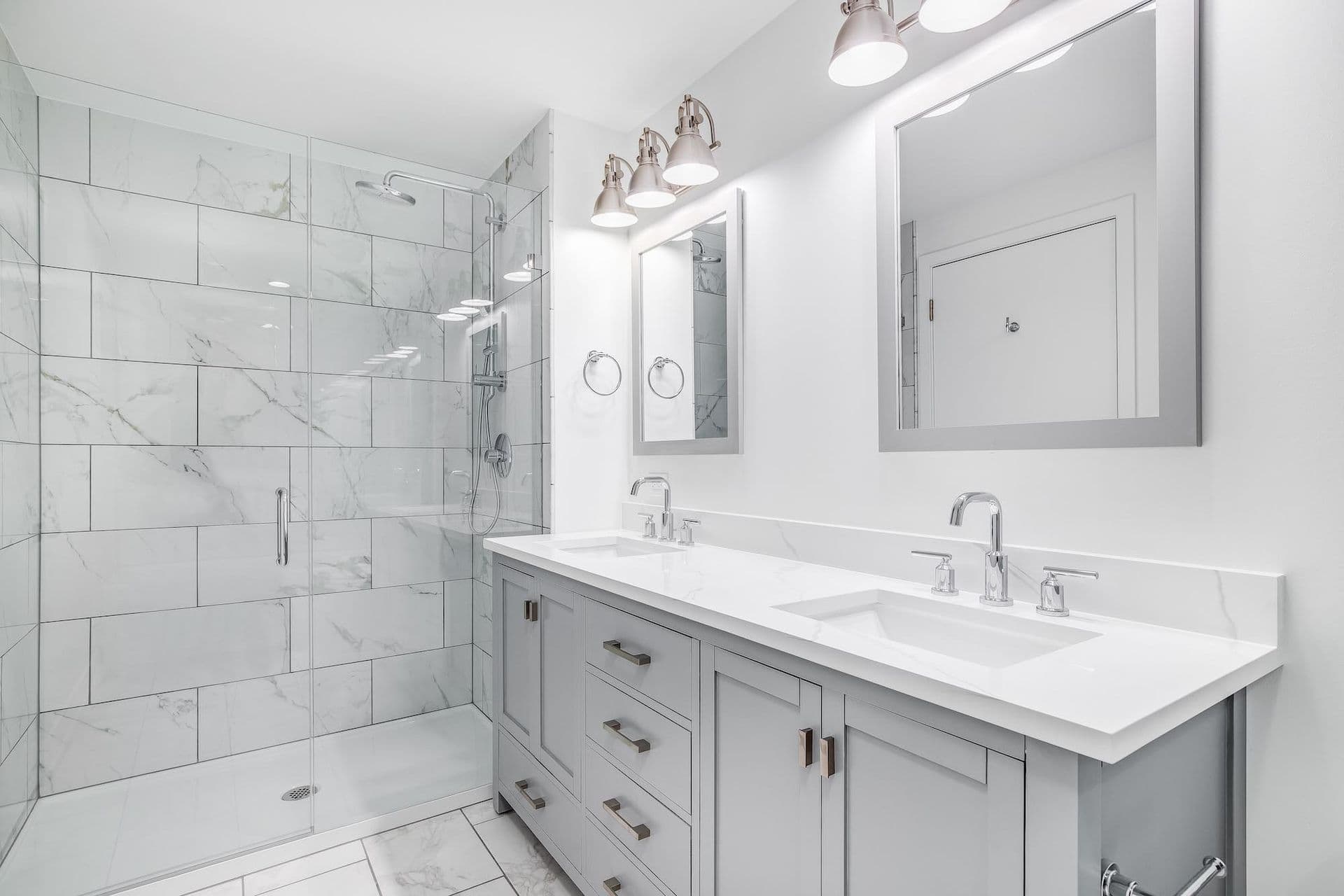 Bright, modern bathroom with a double vanity, sleek faucets, and twin mirrors. A glass-enclosed shower with white marble tiles adds elegance.