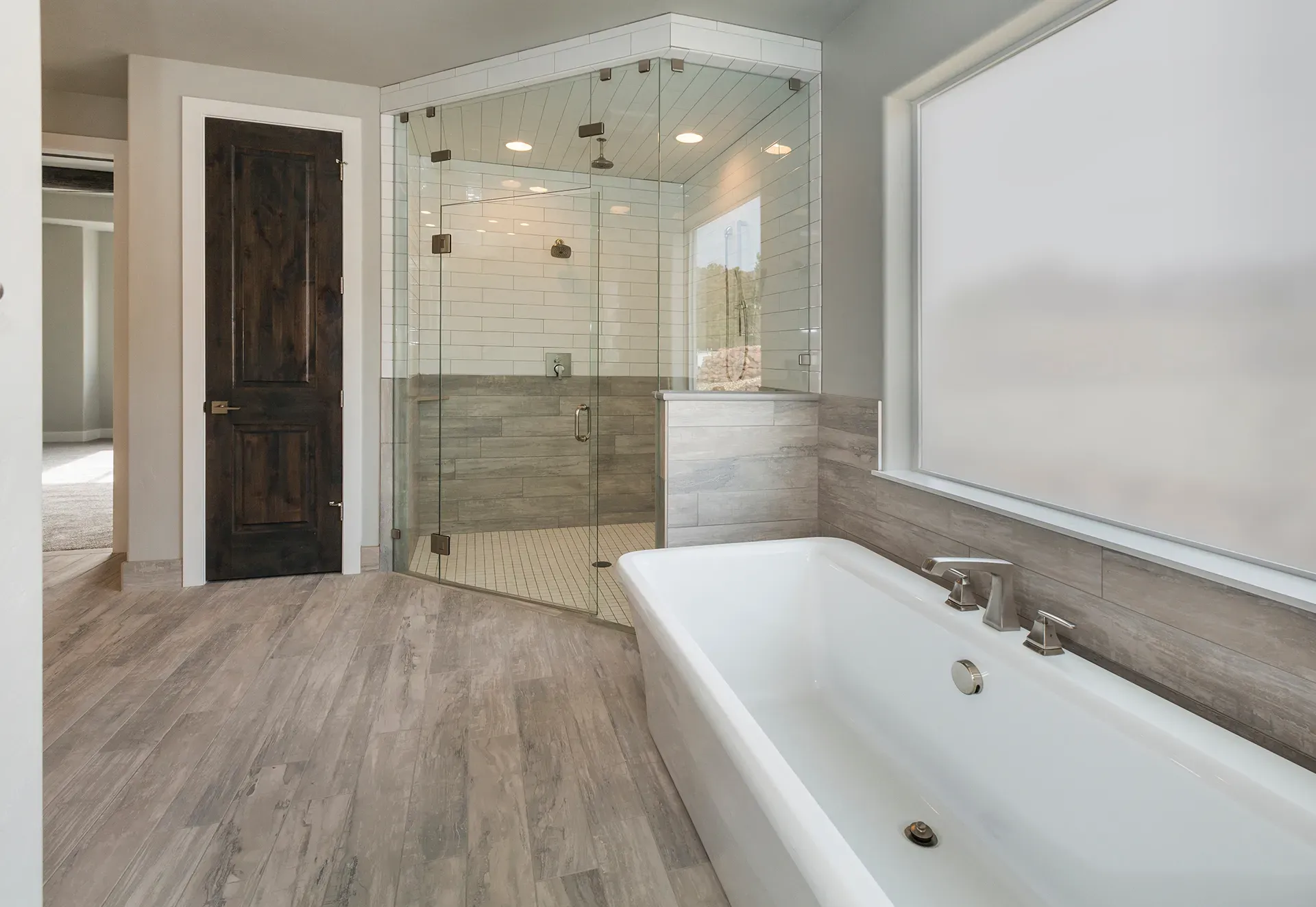 Modern bathroom with a glass-enclosed shower featuring white subway tiles, a white freestanding tub, wood accents, and soft natural light.