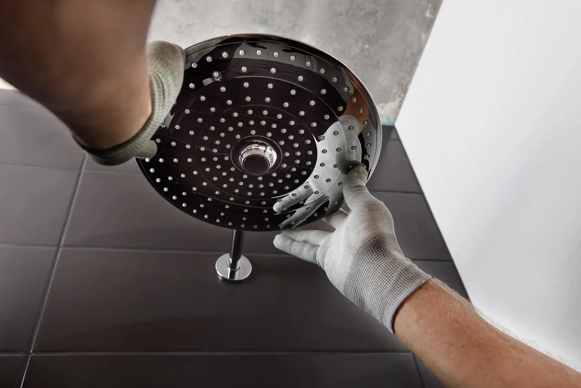 Hands in gray gloves are installing a large, round showerhead with multiple water jets on a tiled bathroom wall.
