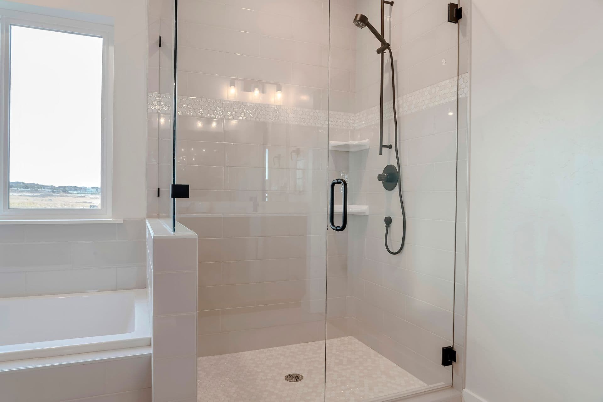 Minimalist bathroom with glass-enclosed shower, white tiles, dark fixtures, and a large window illuminating the space with natural light.