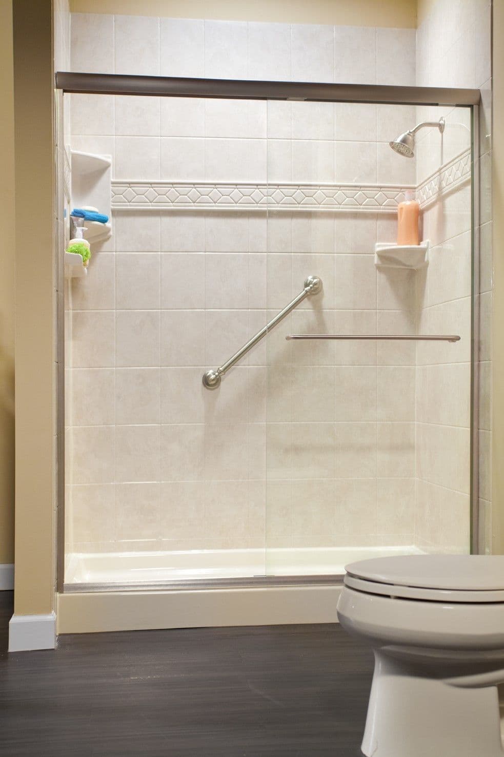Modern bathroom with a glass-enclosed shower featuring cream tiles, a chrome grab bar, and cornershelves. Dark wood floor extends from shower to white toilet.